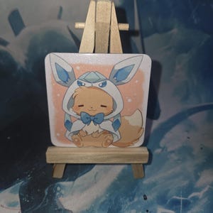 Op de afbeelding: Een vierkante sticker met een stripfiguur in een blauw en wit gehaakt outfit met een blauwe strik. De sticker wordt gepresenteerd op een kleine houten ezel tegen een blauw-witte achtergrond.