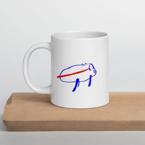 May include: White ceramic mug featuring a blue and red cartoon illustration. The design depicts a stylized animal with a red line across its body and blue legs. The mug is placed on a wooden surface.