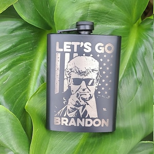 May include: Black metal flask with a silver engraved image of a man wearing sunglasses and a suit jacket. The flask has the text "Let's Go Brandon" on it.