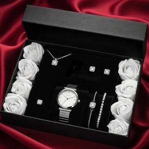 Può includere: Una scatola regalo nera con coperchio aperto, foderata in velluto nero, piena di un orologio argentato, gioielli e rose bianche. I gioielli includono una collana, orecchini, un anello e braccialetti. La scatola è su raso rosso.
