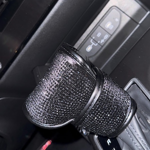 Peut inclure: Un couvre-levier de vitesse de voiture noir et orné de strass. Le couvercle est incrusté de petits strass étincelants et est enveloppé d'une bande de cuir noir. Le levier de vitesse est en position « D ».