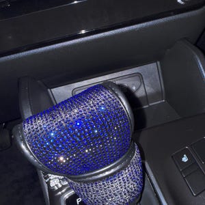 May include: A car gear shift knob cover, encrusted with sparkling blue rhinestones. The cover is black around the edges and fits over the gear shift lever. The background shows the car's console with the gear shift indicator.