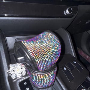 Peut inclure: Un couvre-levier de vitesse et un porte-gobelet de voiture ornés de strass irisés. Les accessoires sont noirs avec une multitude de pierres précieuses colorées. Une figurine Hello Kitty recouverte de strass est également visible.