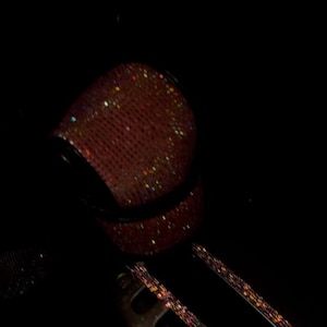 May include: A car gear shift knob and surrounding trim are covered in sparkling, reddish-pink rhinestones. The shift lever is silver with the letters R, N, and D visible.