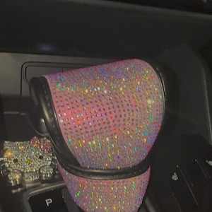 May include: Pink, sparkly car gear shift cover with a black leather trim. The cover is covered in iridescent rhinestones. A silver Hello Kitty figurine is visible in the background.