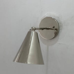 Op de afbeelding: Zilverkleurige wandlamp met een conische kap en een cirkelvormige achterplaat. De lamp is gemaakt van metaal en heeft een gepolijste afwerking. De kap is naar beneden gericht en de lamp is aan een witte muur bevestigd.