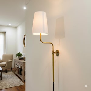 May include: A wall-mounted lamp featuring a white lampshade and a gold-coloured metal frame. The lamp has a curved arm extending from the wall mount to hold the shade. The lamp is against a white wall, and the cord hangs down.