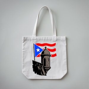 May include: White tote bag featuring a graphic of the Puerto Rican flag and a stylized fort. The flag has a blue triangle with a white star, and red and white stripes. The fort is brown and gray.