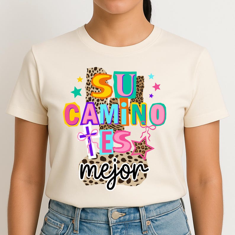 Peut inclure: T-shirt cr&egrave;me avec l'expression espagnole "Su Camino Es Mejor" en lettres color&eacute;es et ludiques. Le motif comprend des imprim&eacute;s l&eacute;opard, des &eacute;toiles et une croix, cr&eacute;ant un graphisme vibrant et accrocheur.