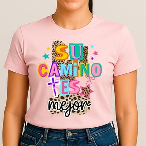 Peut inclure: Un t-shirt rose clair avec l'inscription "SU CAMINO ES MEJOR" en lettres color&eacute;es. Le motif comprend des imprim&eacute;s l&eacute;opard, des &eacute;toiles et une croix. Le t-shirt est fait d'une mati&egrave;re douce.