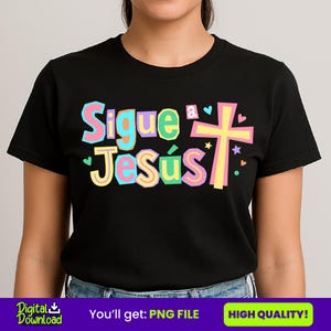 May include: Black crew neck t-shirt with the text "Sigue a Jesús" in colourful letters, a cross, hearts, and stars. The shirt is made of a soft material.