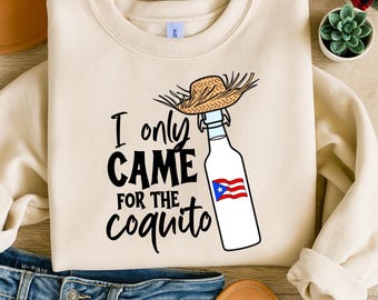 May include: Cream-colored sweatshirt with the text "I only CAME for the coquito." A white bottle with a straw hat on top and the Puerto Rican flag is featured in the design. The sweatshirt is displayed on a neutral background.