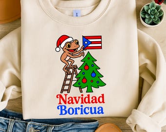 May include: Cream-colored sweatshirt featuring a cartoon frog wearing a Santa hat, placing a Puerto Rican flag on a Christmas tree. The words "Navidad Boricua" are written in red and blue. The design includes a ladder and ornaments.