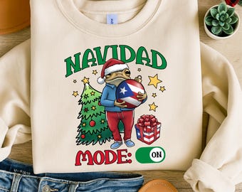 May include: Cream-colored sweatshirt with a Christmas-themed graphic. The design features a frog wearing a Santa hat, holding a Puerto Rican flag ornament, and the text "NAVIDAD MODE: ON". A Christmas tree and gift box are also included.