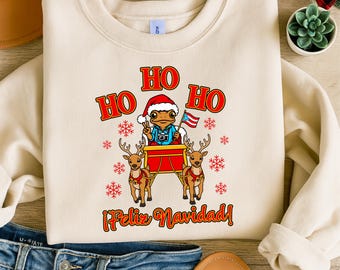May include: A cream-colored sweatshirt with a festive Christmas design. The graphic features a frog wearing a Santa hat and holding a Puerto Rican flag, riding a sleigh pulled by reindeer. The words "HO HO HO" and "¡Feliz Navidad!" are displayed in red.