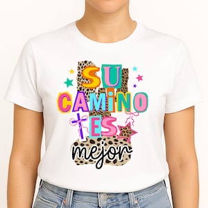 Peut inclure: T-shirt blanc avec l'inscription espagnole "SU CAMINO ES mejor" en lettres color&eacute;es. Le motif comprend des imprim&eacute;s l&eacute;opard, des &eacute;toiles et une croix. Le t-shirt est de style col rond.