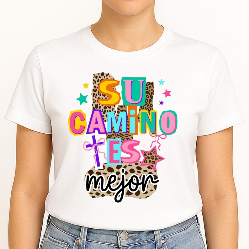 Peut inclure: T-shirt blanc avec l'inscription espagnole "SU CAMINO ES mejor" en lettres color&eacute;es. Le motif comprend des imprim&eacute;s l&eacute;opard, des &eacute;toiles et une croix. Le t-shirt est de style col rond.
