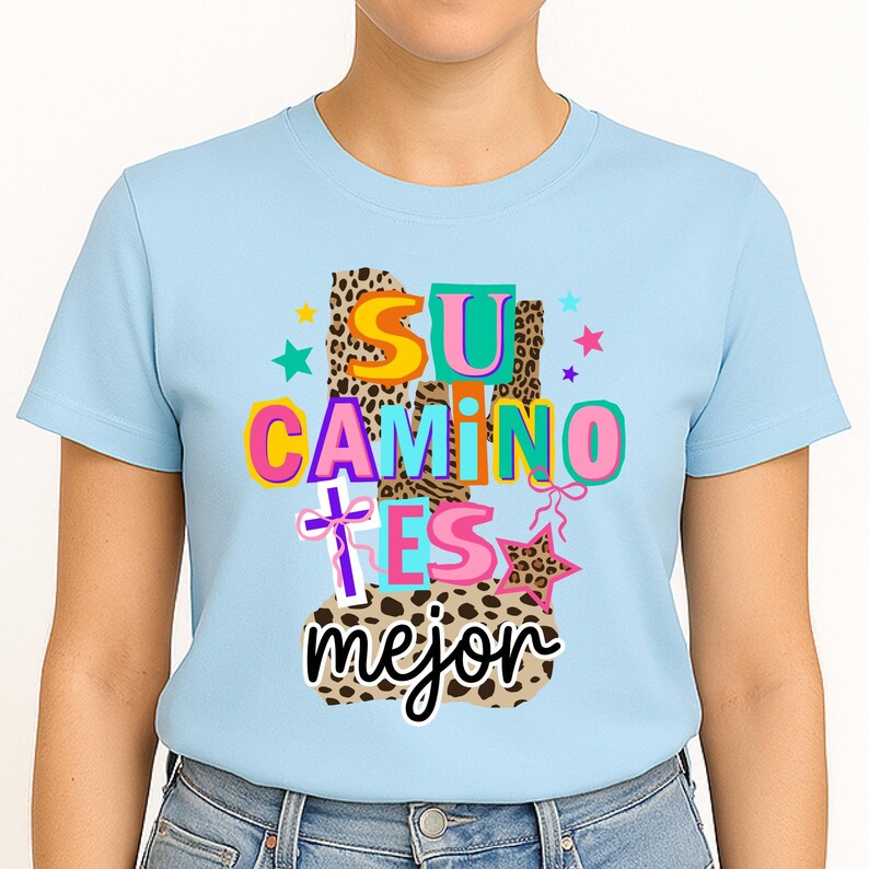 Peut inclure: T-shirt bleu clair avec l'inscription espagnole "SU CAMINO ES mejor" dans une typographie color&eacute;e et ludique. Le motif comprend des &eacute;toiles, une croix et un imprim&eacute; l&eacute;opard, cr&eacute;ant une esth&eacute;tique joyeuse et vibrante.