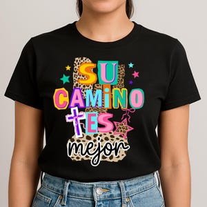Peut inclure: T-shirt noir avec l'inscription color&eacute;e "SU CAMINO ES MEJOR". Le design comprend des motifs l&eacute;opard, des &eacute;toiles et une croix, cr&eacute;ant un graphisme vibrant et accrocheur.