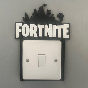May include: A white light switch cover with the word "FORTNITE" in white block letters. The top of the cover has a black silhouette of a landscape. The light switch is in the centre of the cover.