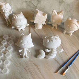 May include: A selection of unpainted white cat figurines, including a mermaid cat and a cat in a cupcake. Also included are paint pots and two paint brushes with blue handles. Perfect for a creative painting project.
