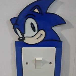 May include: A blue and white Sonic the Hedgehog light switch cover. The cover features a smiling Sonic head above a white light switch set in a blue frame. The design is mounted on a white wall.