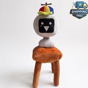 May include: A plush toy featuring a light gray, rounded head with a black screen-like face, white pixelated eyes, and a triangular nose. The toy wears a colorful propeller beanie with red, blue, and yellow panels, and sits on a brown plush stool.
