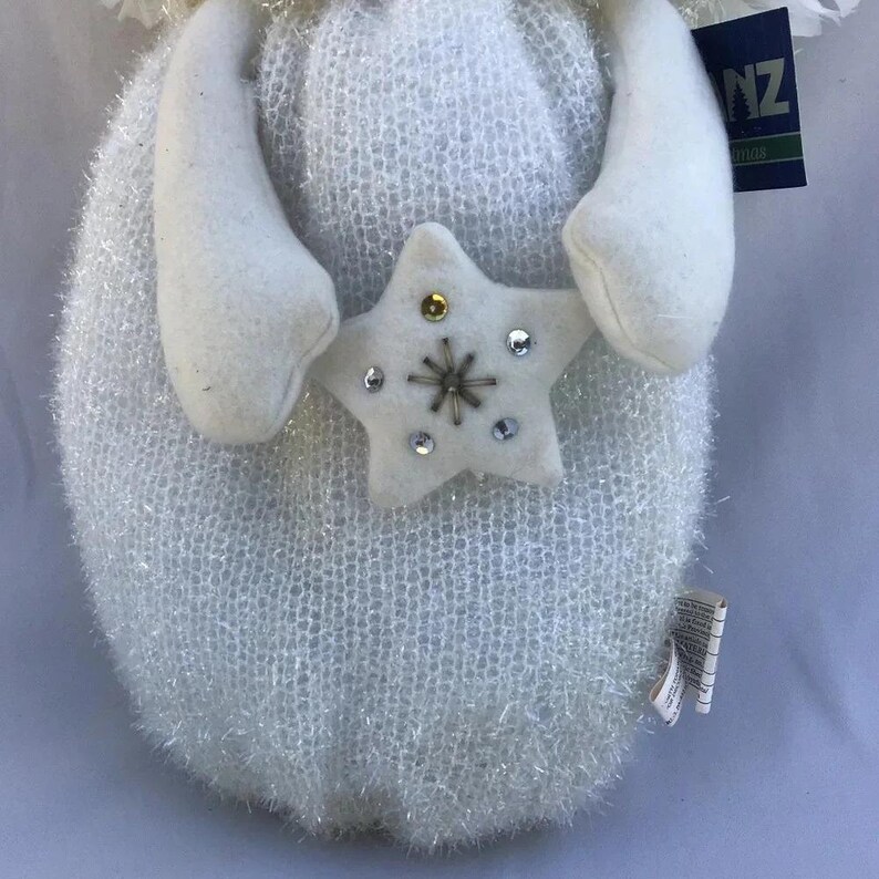 May include: A white angel ornament with a star-shaped decoration. The star is adorned with small, sparkling gems and a gold centre. The angel has white, felt-like arms and a textured body. A tag with the letters "NZ" is visible.