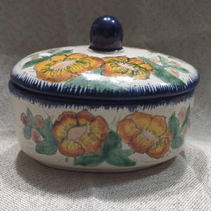 以下が含まれることがあります： 手描きの花柄が施された、蓋付きのセラミック容器。容器はオフホワイトで、オレンジと黄色の花、緑の葉、ネイビーブルーの縁と蓋が付いています。蓋にはネイビーブルーのつまみが付いています。