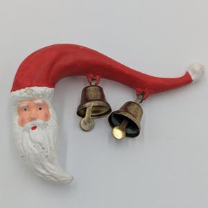 Puede incluir: Un adorno navideño caprichoso con Papá Noel, un gorro rojo y barba blanca. Dos pequeñas campanas de latón cuelgan del gorro. El adorno está pintado con una estética vintage.