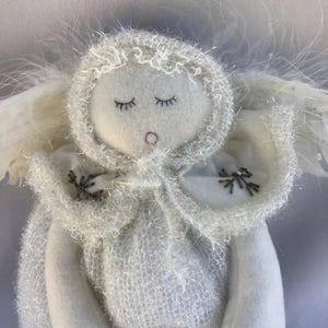 May include: A handmade white angel doll with closed eyes and a small red mouth. The doll has a fluffy, textured hood and cape, with delicate feather wings. The angel's cape is adorned with small, decorative embellishments.