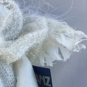 May include: Close-up of a white angel figurine with feathery wings. The angel's wings are made of delicate, layered feathers. The figurine has a soft, textured appearance. The tag on the figurine reads "ANZ".