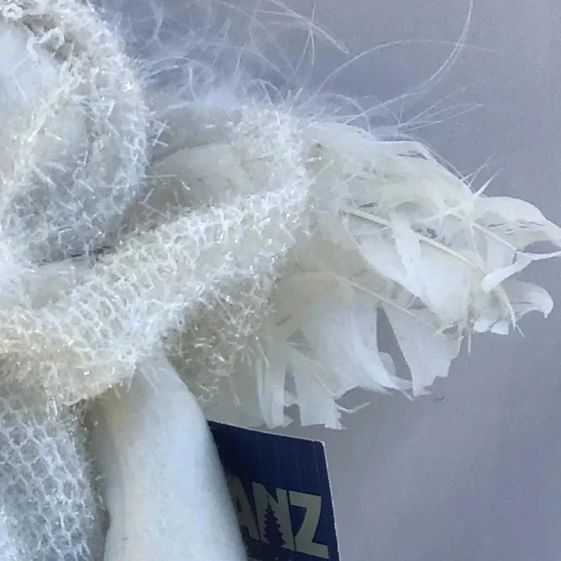 May include: Close-up of a white angel figurine with feathery wings. The angel's wings are made of delicate, layered feathers. The figurine has a soft, textured appearance. The tag on the figurine reads "ANZ".