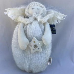 May include: A white angel doll with closed eyes, holding a star. The doll has a fluffy, textured body, arms, and head covering. White feathers form the wings. The star is decorated with small, colourful embellishments. A tag is attached to the doll.