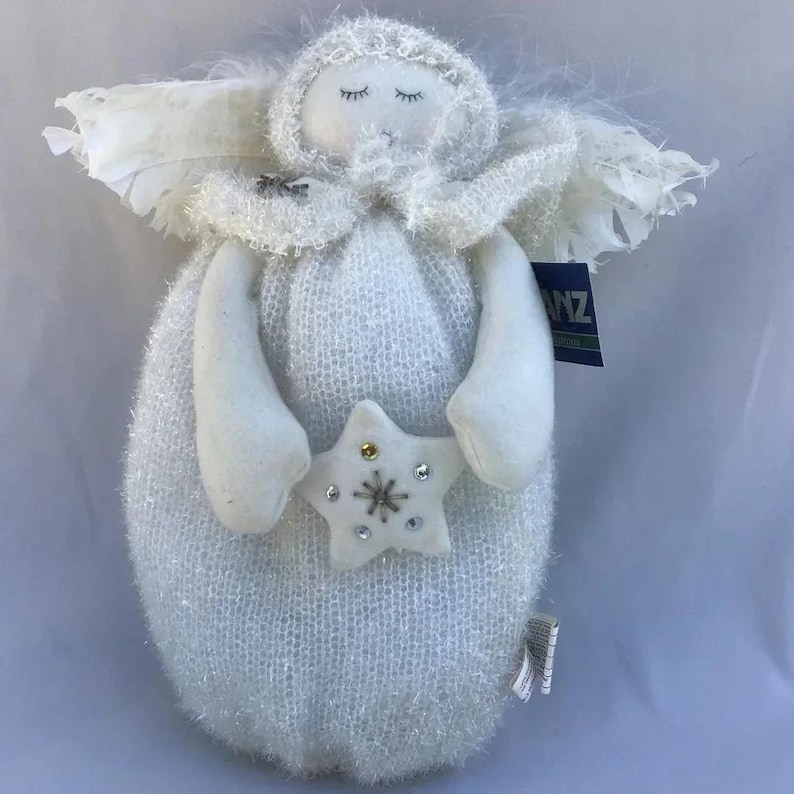 May include: A white angel doll with closed eyes, holding a star. The doll has a fluffy, textured body, arms, and head covering. White feathers form the wings. The star is decorated with small, colourful embellishments. A tag is attached to the doll.