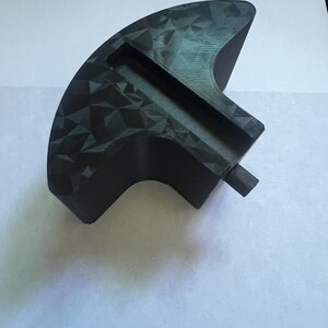 May include: A black, geometric-patterned object with a curved shape and a rectangular slot. The object has a textured surface and a small, cylindrical protrusion. The item is likely a tool or a component of a larger device.