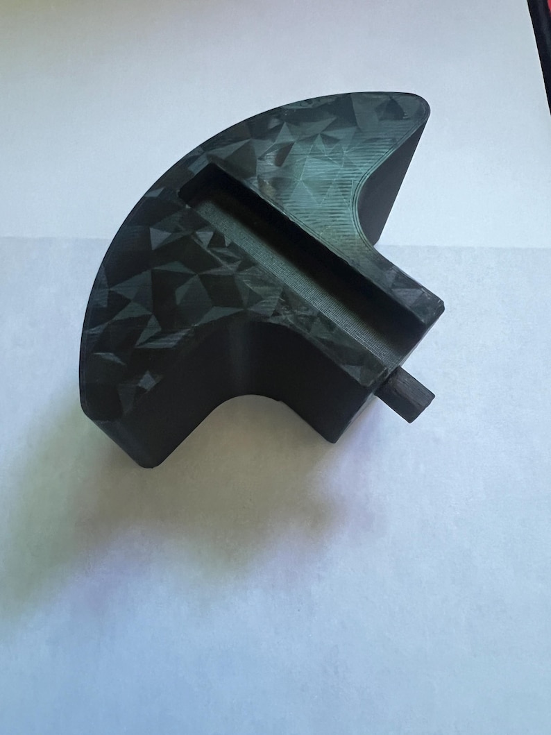 May include: A black, geometric-patterned object with a curved shape and a rectangular slot. The object has a textured surface and a small, cylindrical protrusion. The item is likely a tool or a component of a larger device.