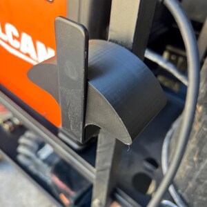 May include: Close-up of a black, curved plastic component, possibly a bracket or holder, attached to a larger object. The component has a textured surface and is positioned near an orange and black object with the number "220" visible.