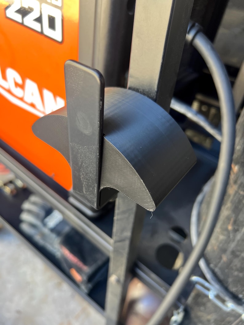 May include: Close-up of a black, curved plastic component, possibly a bracket or holder, attached to a larger object. The component has a textured surface and is positioned near an orange and black object with the number "220" visible.