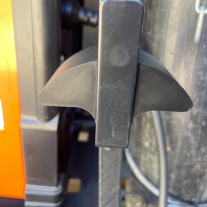 May include: A close-up of a black lever with a textured, wing-like design. The lever is attached to a larger, dark-colored machine with an orange accent. The background includes a metal cylinder and a chain.