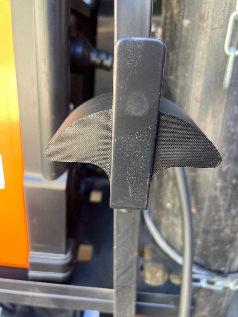 May include: A close-up of a black lever with a textured, wing-like design. The lever is attached to a larger, dark-colored machine with an orange accent. The background includes a metal cylinder and a chain.