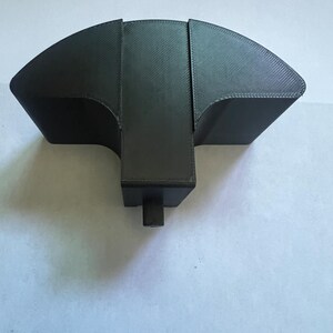 May include: A black, 3D-printed object with a unique shape. It features a curved, fan-like top section and a rectangular base with a cylindrical projection. The object appears to be made of a textured material.