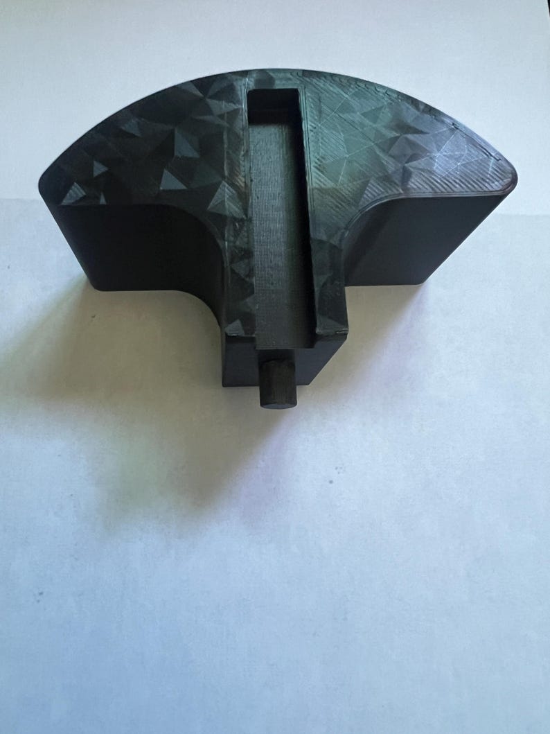 May include: A black, geometric-patterned object with a fan-like shape. It features a central rectangular channel and a cylindrical projection at the base. The surface has a textured, triangular design.