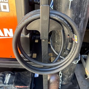 May include: A black welding machine cart with a coiled black cable, a black glove, and a brown metal object. The cart has an orange panel with white text. A black wheel is visible at the bottom.