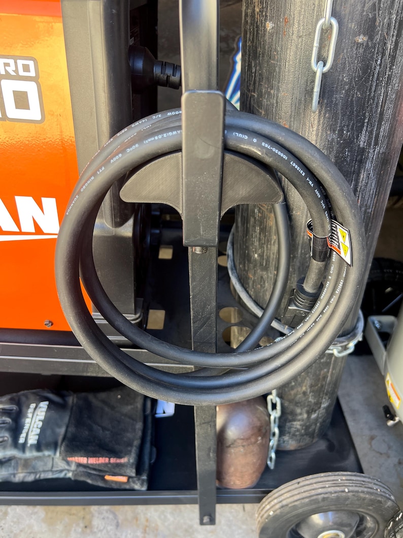 May include: A black welding machine cart with a coiled black cable, a black glove, and a brown metal object. The cart has an orange panel with white text. A black wheel is visible at the bottom.