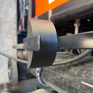May include: A black, curved metal component attached to a machine. The component is cylindrical and appears to be made of a solid material. The background includes parts of the machine, including an orange panel and a wheel.
