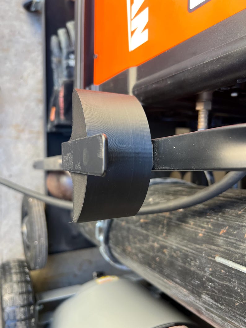 May include: A black, curved metal component attached to a machine. The component is cylindrical and appears to be made of a solid material. The background includes parts of the machine, including an orange panel and a wheel.