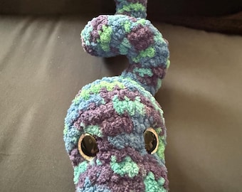 May include: A handmade, plush snake toy in shades of blue, green, and purple. The snake has large, round, black eyes and is made of a soft, textured yarn. The toy is designed with a curved body.