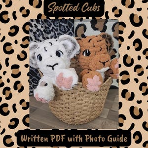 May include: Two crocheted lion cubs, one white and one brown, sit in a woven basket. The cubs have pink paw pads and are labeled "Spotted Cubs" and "Written PDF with Photo Guide". The background features a leopard print.