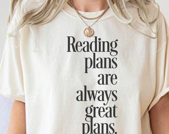 May include: Cream-colored t-shirt with the text "Reading plans are always great plans" in black, bold font. The shirt has short sleeves and a relaxed fit. A gold necklace with a pendant is visible.